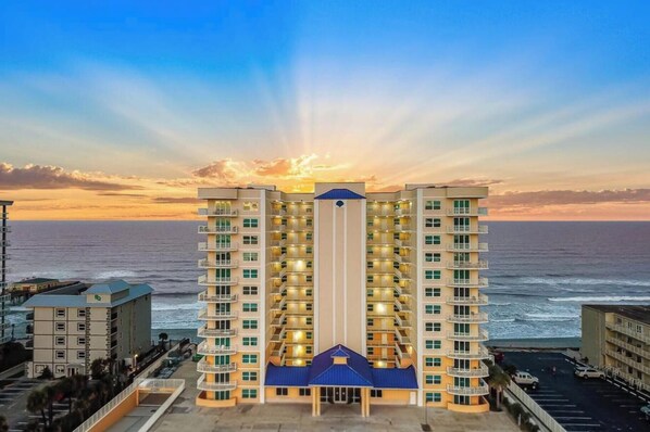 A multi-story hotel with balconies, a central blue-roofed structure, and a view of the ocean. - Spacious Upscale 3BR Condo Balcony Ocean View & Steps to Beach (Daytona Beach)