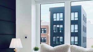 A bedroom with a bed, a nightstand with a lamp, a window, and a view of buildings.