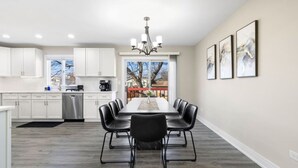 A modern kitchen with white cabinets, a dining area with a long table and black chairs, and a chandelier. - Finally… A Home Big Enough for Everyone 8 Beds (Woodstock)