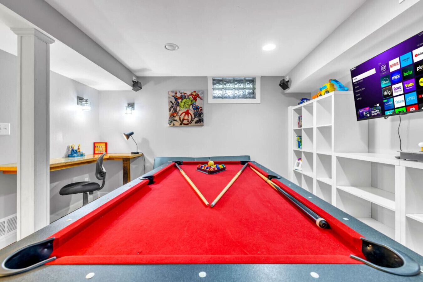 Ein Billardtisch mit roter Spielbahn, ein Billardstange und ein farbenfroher Billardkugelständer. Außerdem ein Bücherregal mit diversen Gegenständen und ein an der Wand montierter Fernseher, der eine Streaming-Dienstanwendung zeigt.