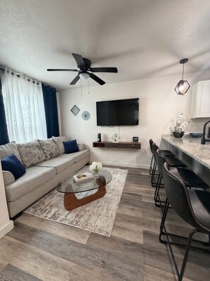 Ein modernes Wohnzimmer mit Sofa, Couchtisch, Fernseher, Deckenventilator und Küchenzeile mit Barhocker.