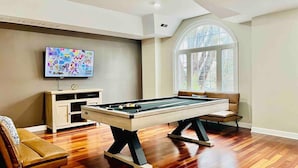 Une table de billard dans une pièce dotée d’un plancher en bois, d’un canapé, d’un téléviseur et d’une fenêtre donnant sur des arbres.