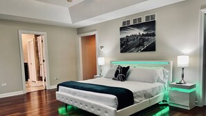 Une chambre moderne avec un grand lit, un tableau représentant un paysage urbain et un éclairage vert placé sous le lit.