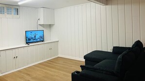 Ein Zimmer mit weiß gepaneltem Mauerwerk, einem Fernseher auf einem Standfuß, einem dunkelgrauen Sofa und Holzboden.