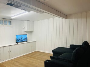 A room with white paneled walls, a television on a stand, a dark-colored sofa, and wooden flooring.