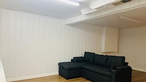 Ein Zimmer mit Ecksofa, Holzboden, weißen Wandverkleidungen und einer Decke mit Einbaustrahlern.