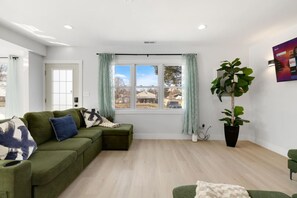 A modern living room with a sectional sofa, a large window with curtains, a potted plant, and a wall-mounted TV. - Elegant 4BR Lakefront Retreat-Ready For Long-Stay (Antioch)