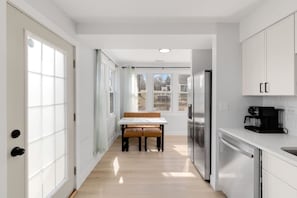 A narrow kitchen with white cabinets, a stainless steel refrigerator, a coffee maker, and a wooden bench at the end. - Elegant 4BR Lakefront Retreat-Ready For Long-Stay (Antioch)