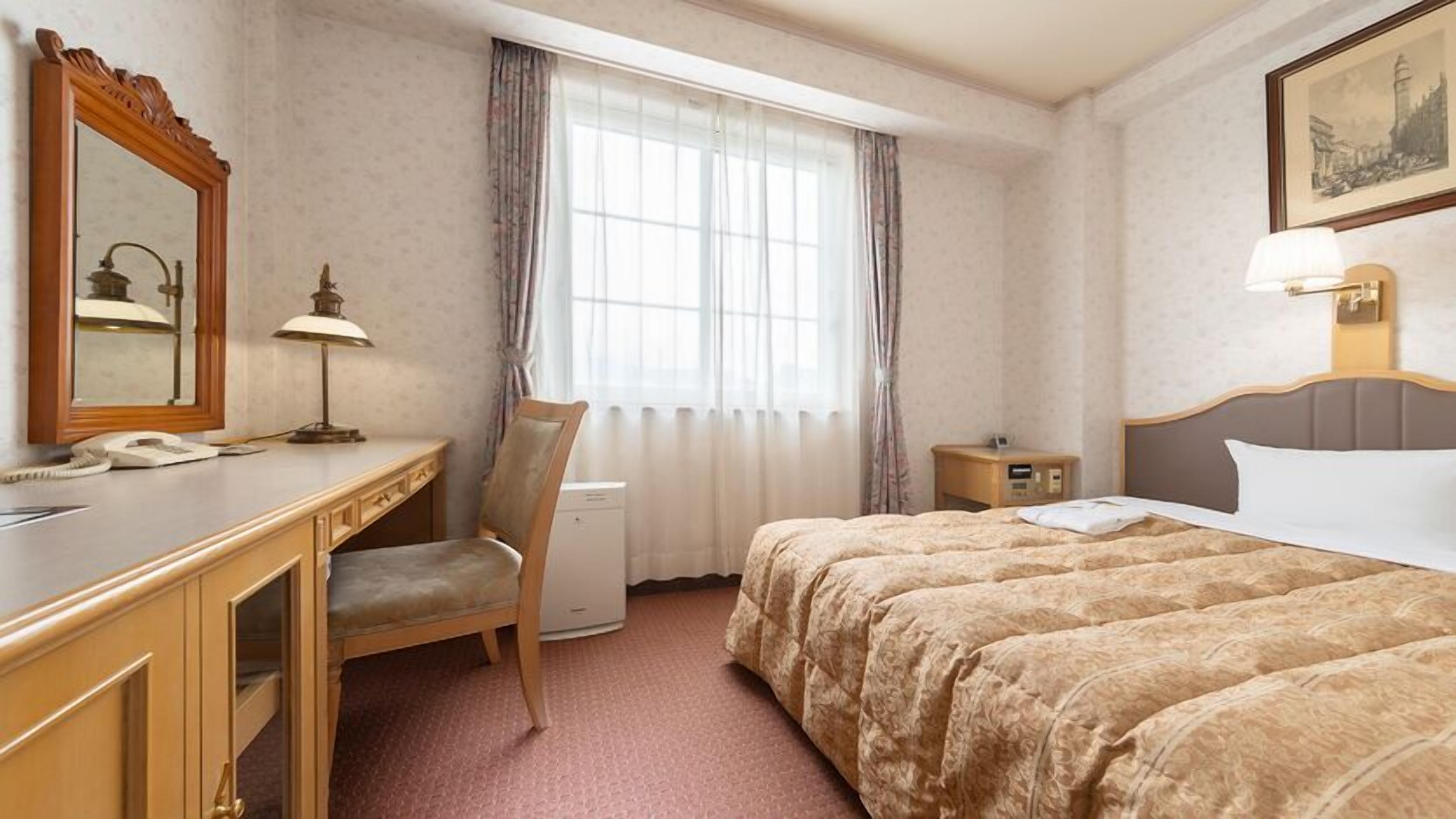 A hotel room with a bed, a desk, a chair, a nightstand, a lamp, a mirror, and a window with curtains.