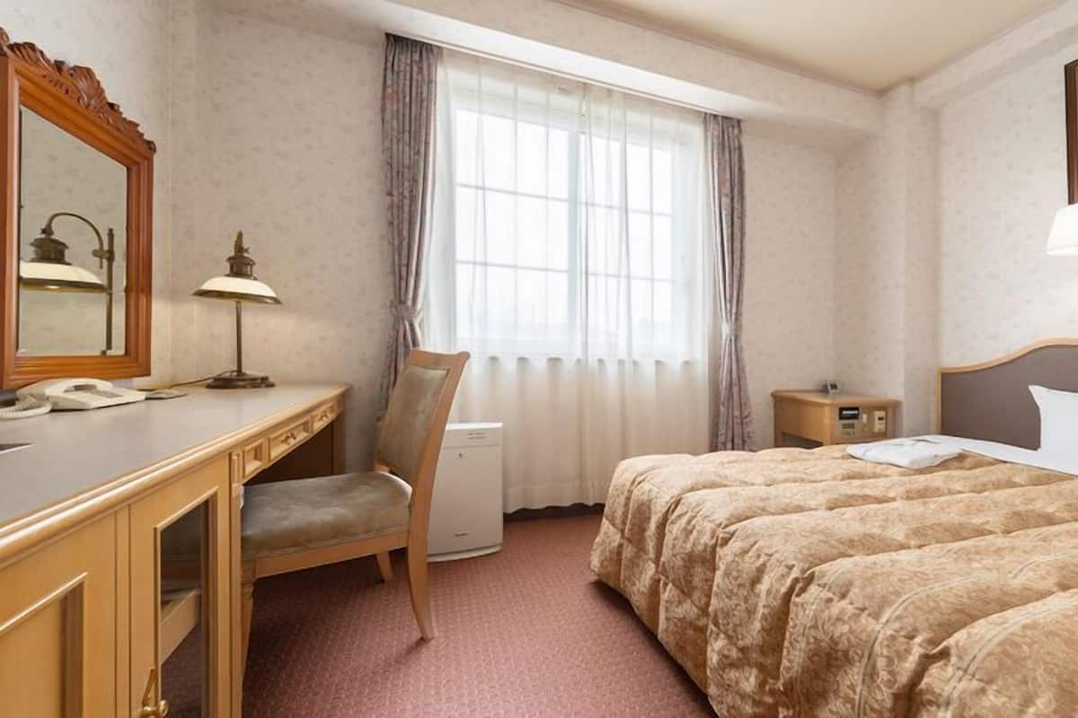 A hotel room with a bed, a desk, a chair, a nightstand, a lamp, a mirror, and a window with curtains.