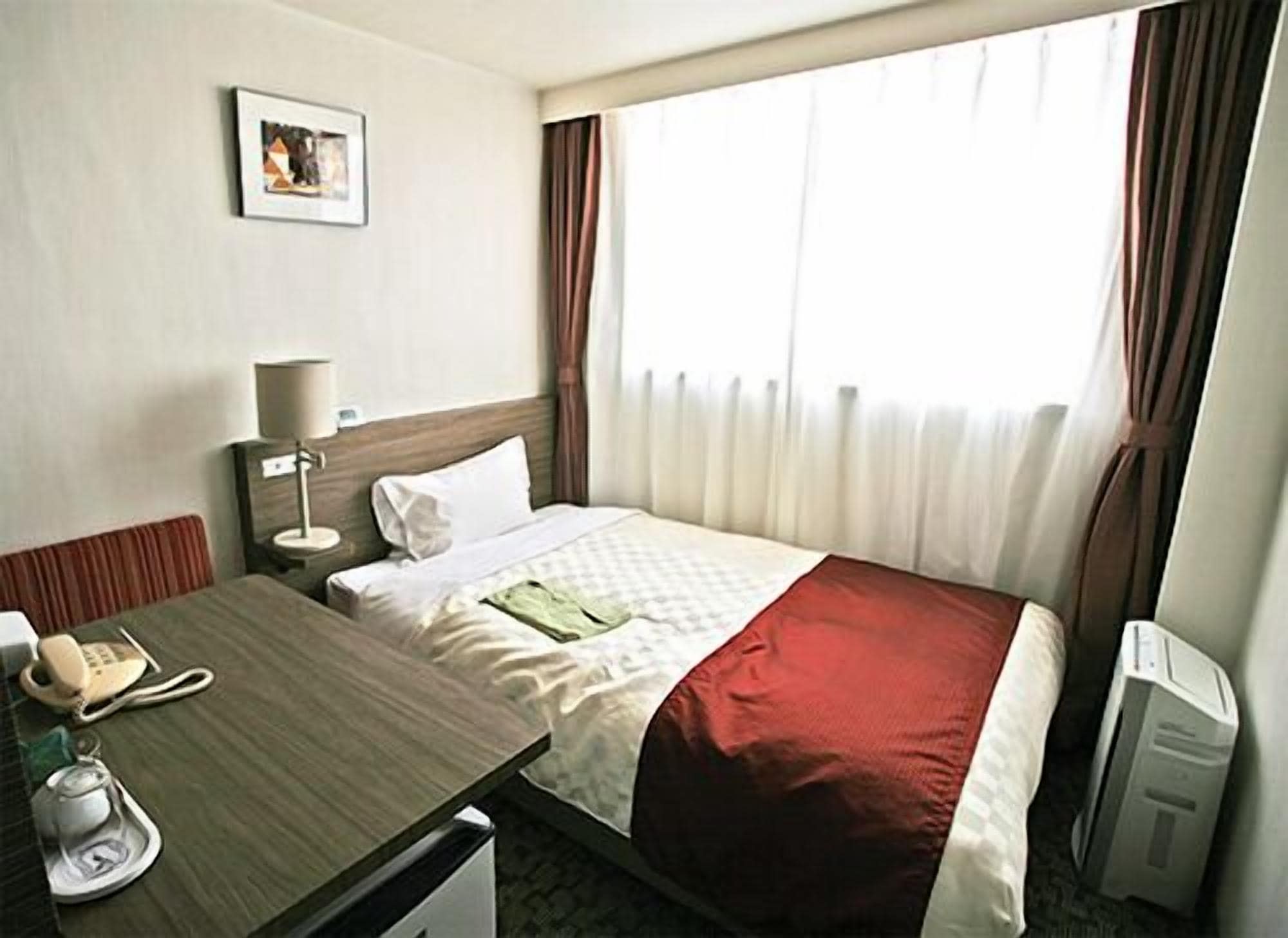 A hotel room with a bed, a desk with a telephone, a lamp, a picture on the wall, and a window with curtains.
