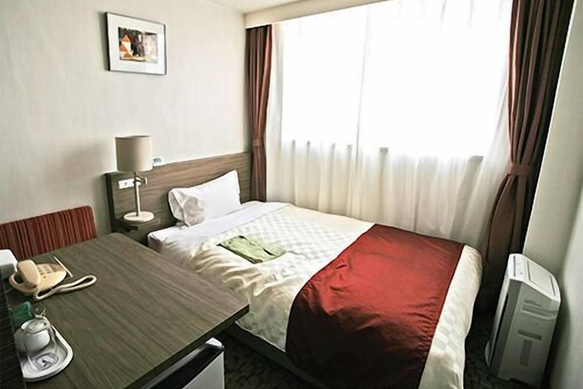 A hotel room with a bed, a desk with a telephone, a lamp, a picture on the wall, and a window with curtains.