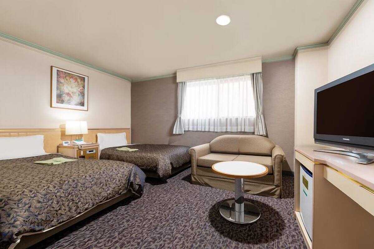 A hotel room with two beds, a sofa, a small round table, a television, and a window with curtains.
