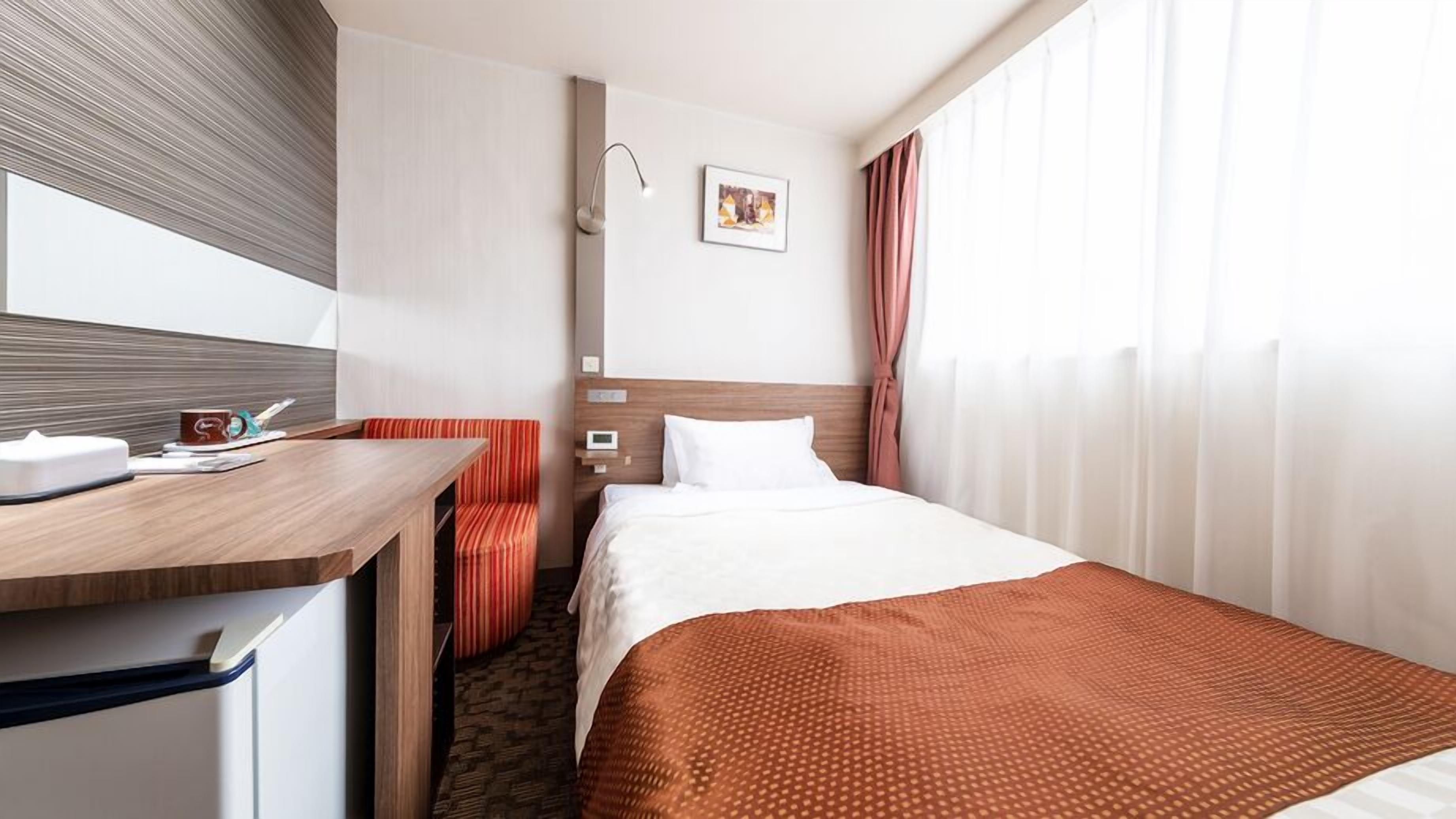 A hotel room with a bed, a desk, a chair, a bedside table, a wall-mounted lamp, a picture, and a window with curtains.