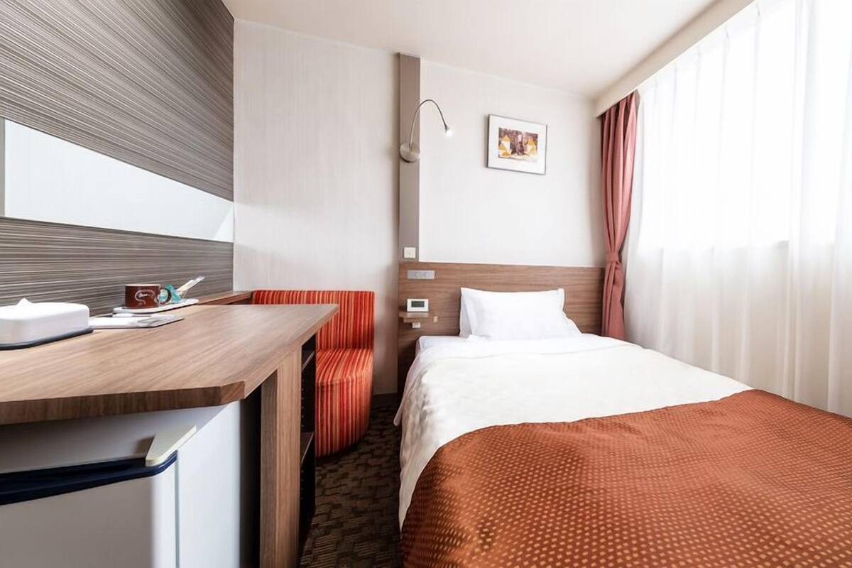A hotel room with a bed, a desk, a chair, a bedside table, a wall-mounted lamp, a picture, and a window with curtains.