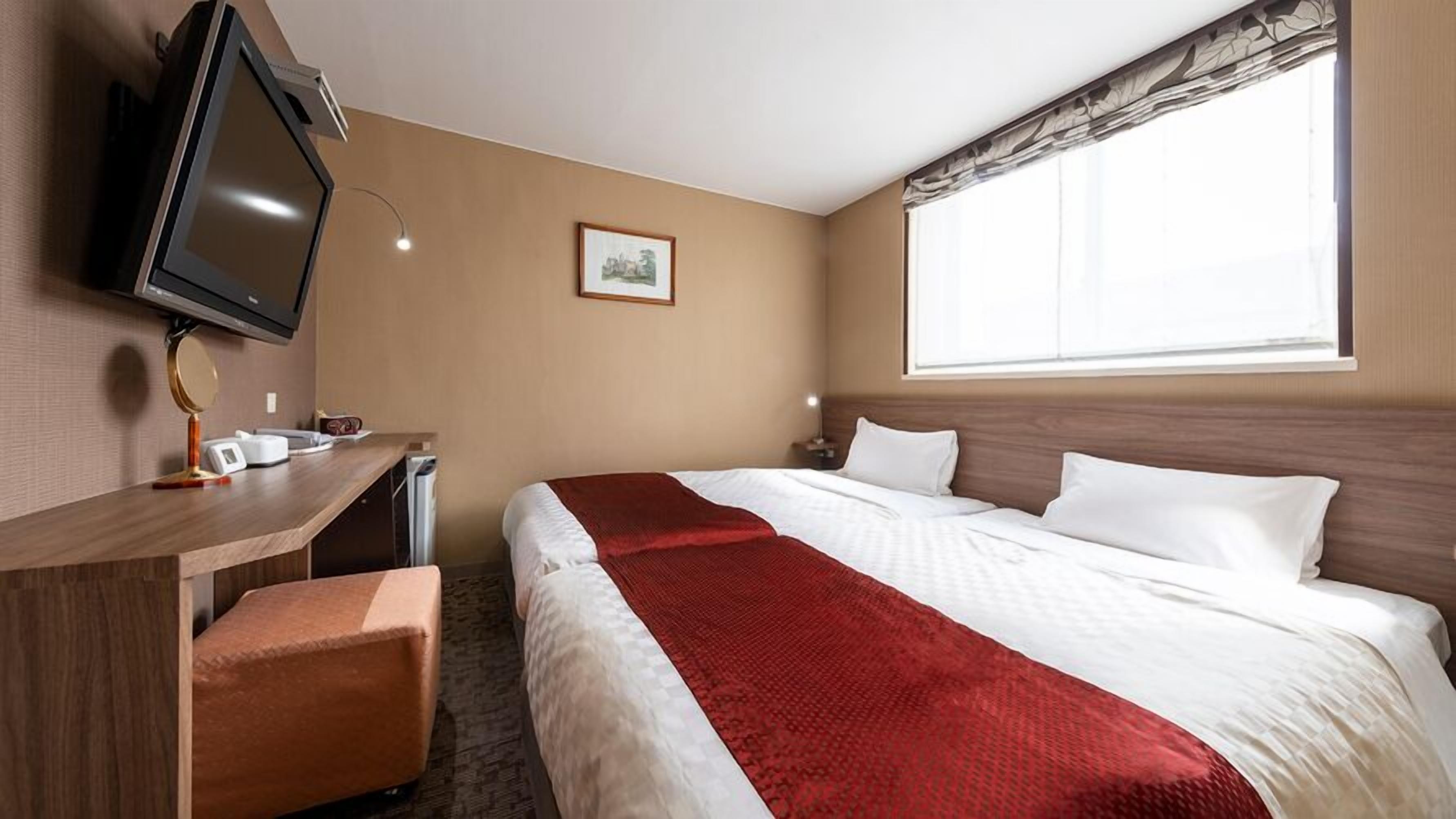 A hotel room with a bed, a desk with a television, a small table, and a window with curtains.