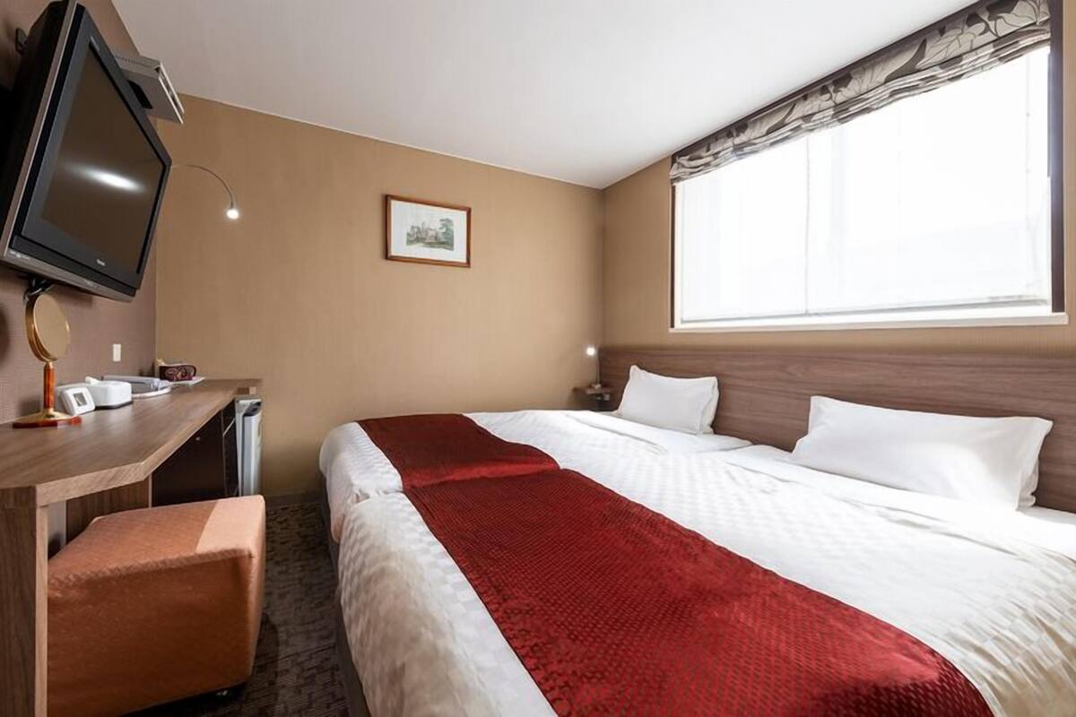 A hotel room with a bed, a desk with a television, a small table, and a window with curtains.