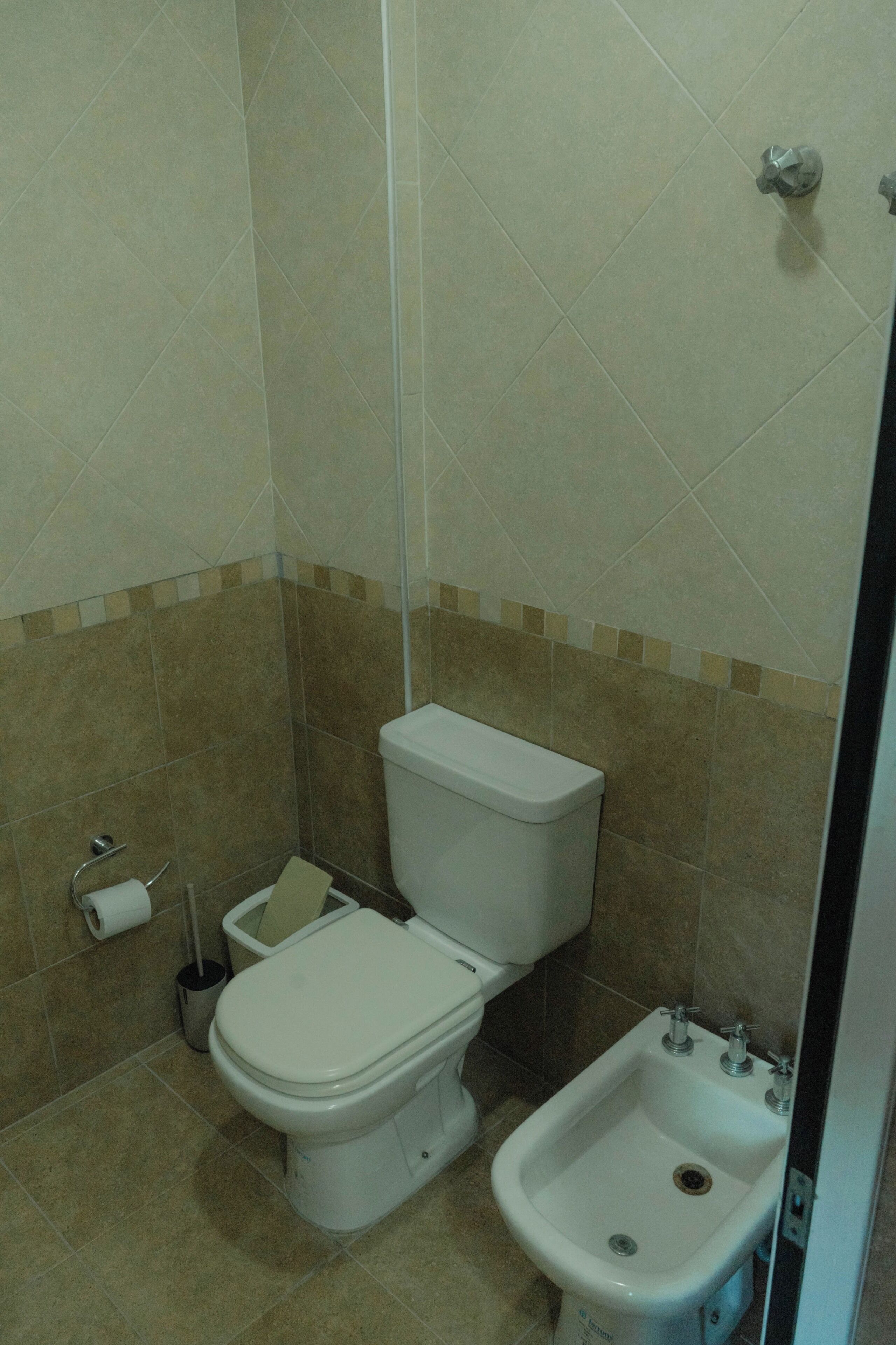 A bathroom with a toilet, bidet, and a roll of toilet paper on the floor.