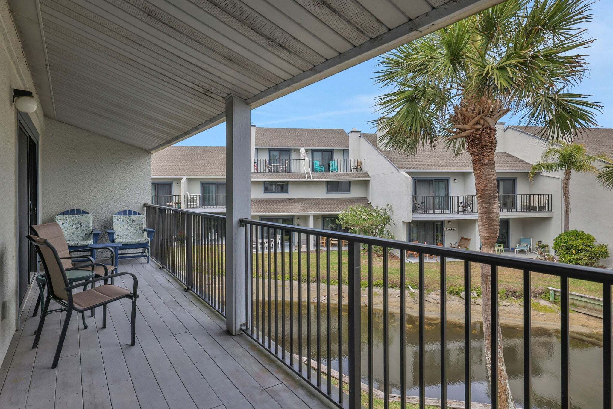 A covered balcony with outdoor furniture, a palm tree, and a view of adjacent buildings.
