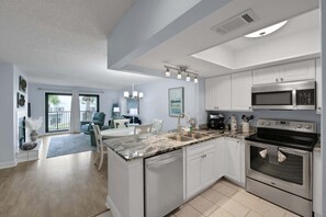 A modern kitchen with white cabinets, a granite countertop, stainless steel appliances, and a dining area with a blue chair. - Sea Winds Hideaway (St. Augustine)