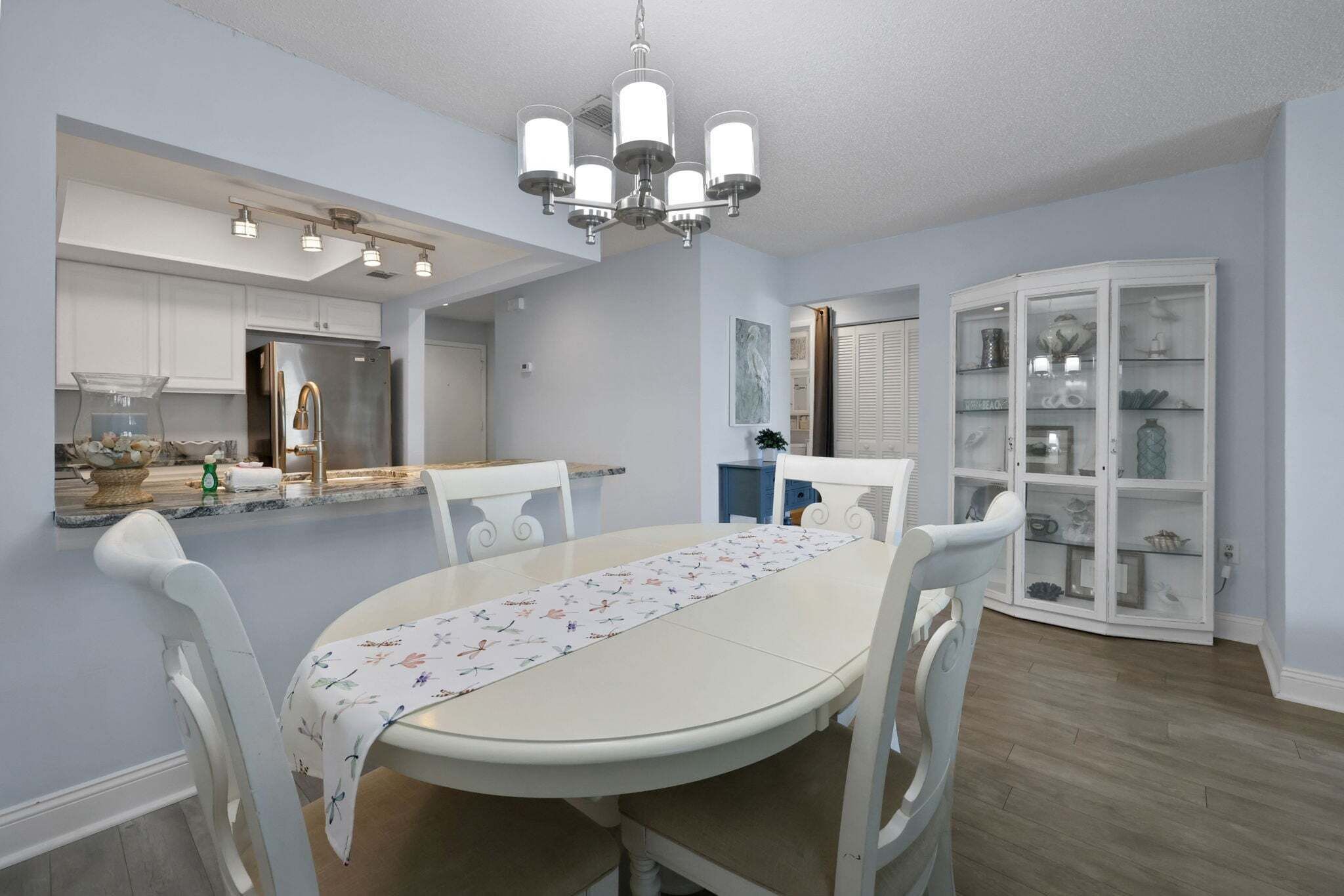A dining area with a white table, chairs, a chandelier, a kitchen in the background, and a glass cabinet with decorative items.