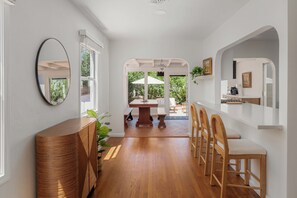 Un pasillo con suelo de madera, un espejo redondo, una zona de comedor con mesa y sillas, y una cocina con mostrador y sillas.