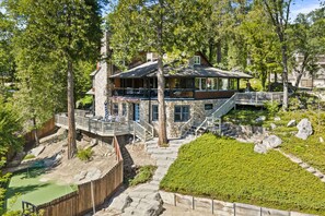 A two-story stone house with a deck, a stone staircase, and a landscaped yard. - Lakeview | Dock Access | Sleeps 16 | Putting Green (Bass Lake)