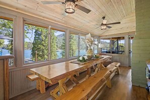 A wooden dining area with a large table and benches, large windows with a view of trees and water, and a ceiling fan. - Lakeview | Dock Access | Sleeps 16 | Putting Green (Bass Lake)
