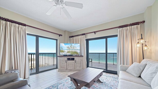 A spacious living area with a flat-screen TV, a wooden coffee table, a sofa, and a balcony with a view of the ocean. - The Palms East 901 | Beachfront | Pools | My Beach Getaways (Orange Beach)