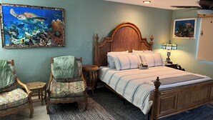 Ein Schlafzimmer mit einem Bett, zwei Stühlen, einem runden Beistelltisch, einem hölzernen Kopfteil, einem Teppich und zwei an der Wand befestigten Bildern.