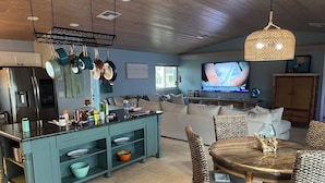 Eine moderne Küche mit Kochinsel, Essbereich mit Korbstühlen und Fernseher im Wohnzimmer.