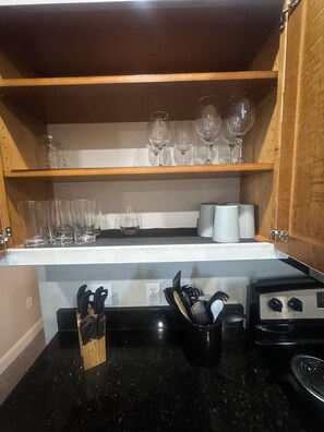 A kitchen with a countertop, a stove, and a cabinet with wine glasses and utensils.