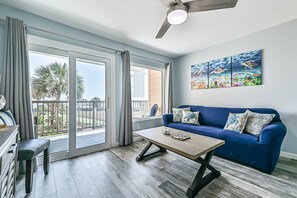 A living room with a blue sofa, a wooden coffee table, a ceiling fan, and a large window with a view of palm trees. - The Turquoise Turtle, Sleeps 4 with Stunning Oceanview. New Pools! (Galveston)
