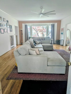 Ein Wohnzimmer mit Ecksofa, Deckenventilator und großem Fenster mit Vorhängen.