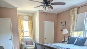 Ein Schlafzimmer mit einem Bett, einem Sessel, einem Schreibtisch, einem Deckenventilator und einem Fenster mit Vorhängen.