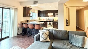A modern living room with a grey sofa, a small ottoman, and a kitchen area with dark cabinets and a central island.