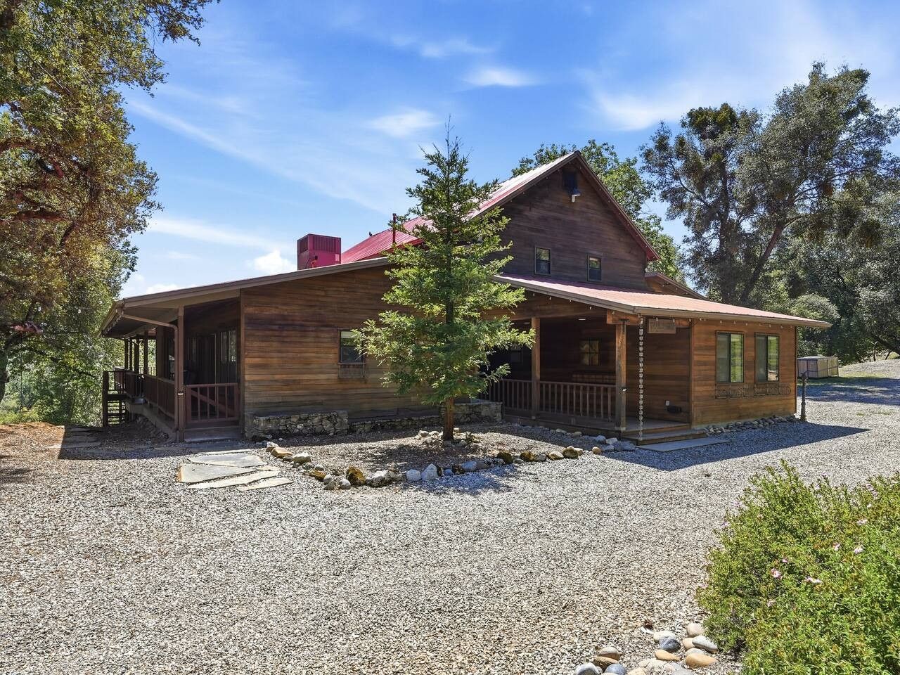 A wooden cabin with a porch, a small tree in front, and a gravel driveway.