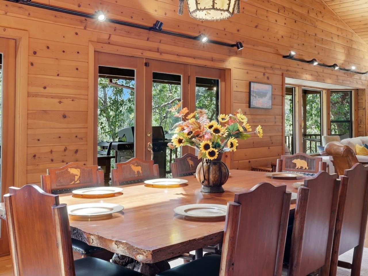 A wooden dining room with a large table and chairs, a vase of sunflowers, and a view of the outdoors.