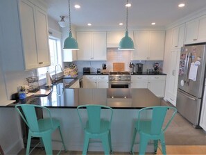 A modern kitchen with white cabinets, a central island, teal pendant lights, and a stainless steel refrigerator. - Ocean View Beach Block Condo | 4BR | Private Elevator | Large Deck | Sleeps 10 (Ocean City)
