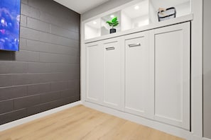 Ein Zimmer mit grauer Ziegelwand, einem weißen Kleiderschrank mit drei Türen, einem kleinen Regal mit einer Pflanze und einem Wand-Fernseher.