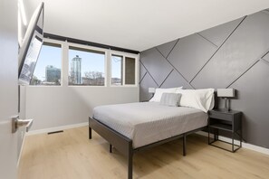 Ein Schlafzimmer mit Bett, Nachttisch, Fenster und Blick auf Gebäude.