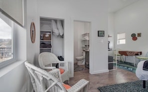 Une pièce avec un fauteuil en osier, une armoire blanche, une petite salle de bain et une vue sur l’extérieur.