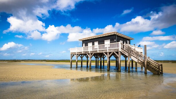 Een houten steiger die leidt naar een klein gebouw met een balkon, gelegen op een zandstrand met heldere lucht.