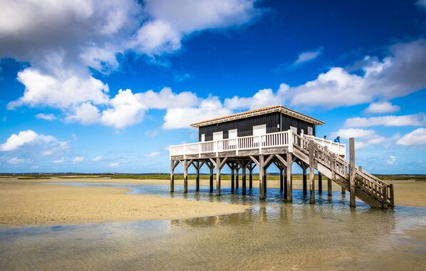 Eine Holzstege führt zu einem kleinen Gebäude mit einem Balkon, gelegen an einem sandigen Strand unter blauem Himmel.