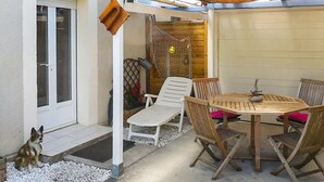 Een terras met een tafel, stoelen, een hond en een overdekt gedeelte met een houten deur.