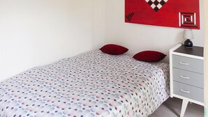 Een slaapkamer met een bed, twee rode kussens, een grijze dressoir met lades, en een abstract schilderij in het rood aan de muur.