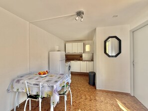 Eine kleine Küche mit weißen Schränken, einem Kühlschrank und einer Waschmaschine. Es gibt eine Essecke mit Tisch und Stühlen sowie einen Mülleimer.