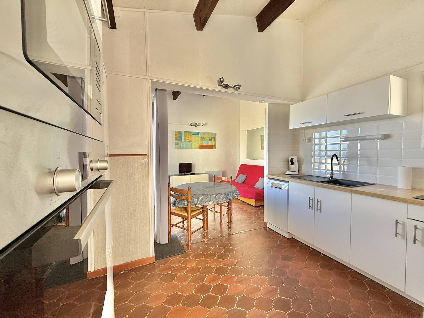 A modern kitchen with white cabinets, a dining area with a red sofa, and a tiled floor.