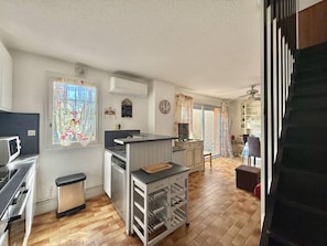 A compact kitchen with a stove, sink, and a small island with a cutting board. There is a window with curtains, a clock, and a ceiling fan. - CAP D'AGDE - MONT ST MARTIN // Réf 0315 (CAP D'AGDE)