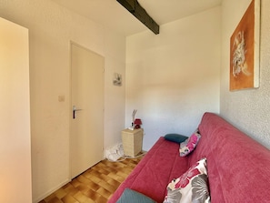 A room with a wooden floor, a red sofa with pillows, a door, and a small table with a lamp. - CAP D'AGDE - MONT ST MARTIN // Réf 0315 (CAP D'AGDE)